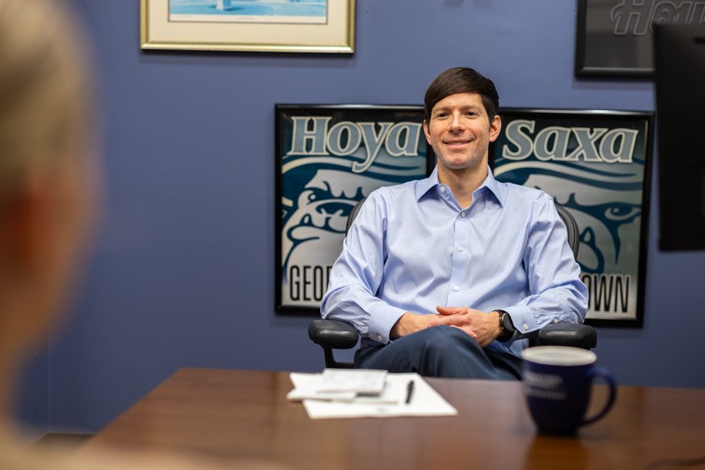 A man smiles from behind a desk at a student in the foreground. He has a coffee mug and legal pad on his desk and behind him are two photos that say "Hoya Saxa"