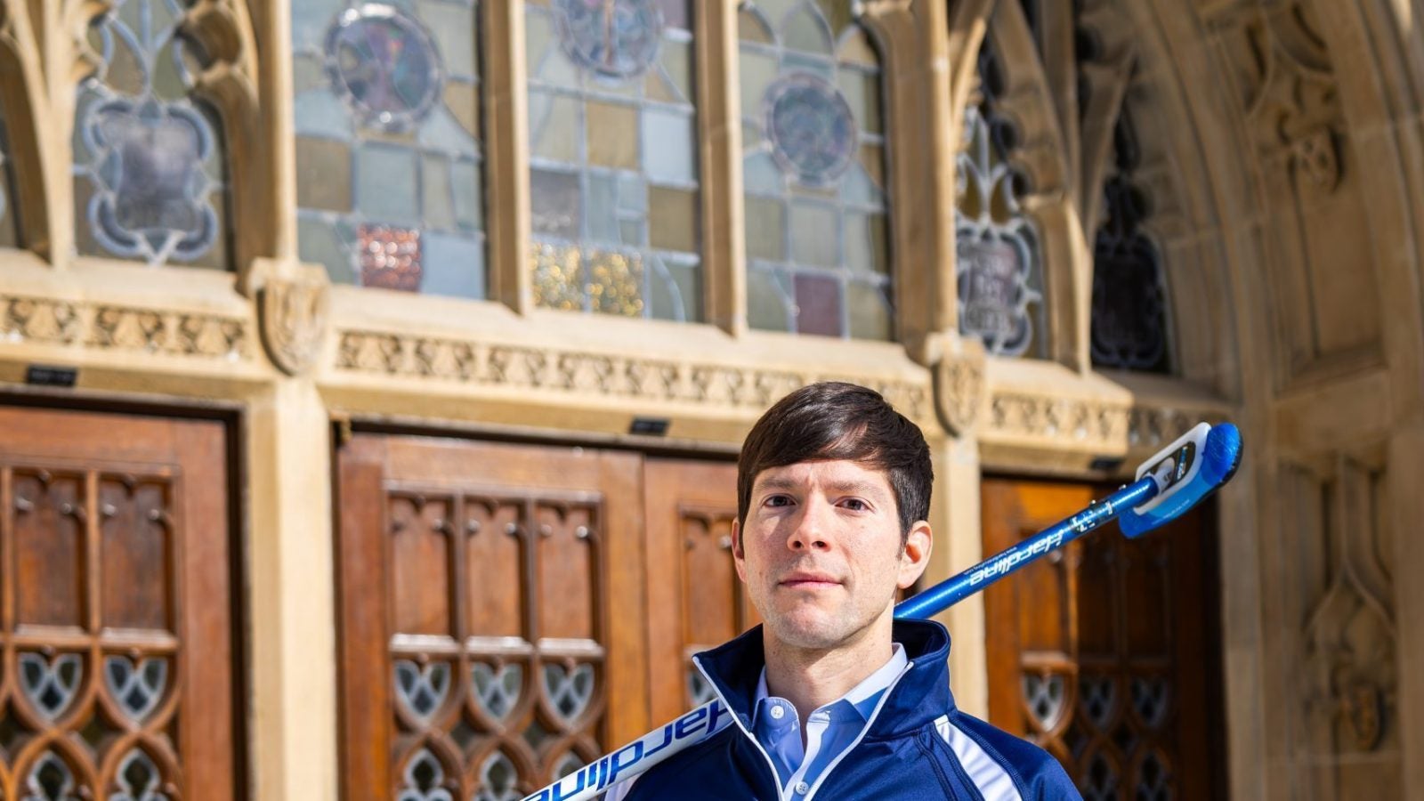 A man with a curling broom stands outside an archway of an academic building