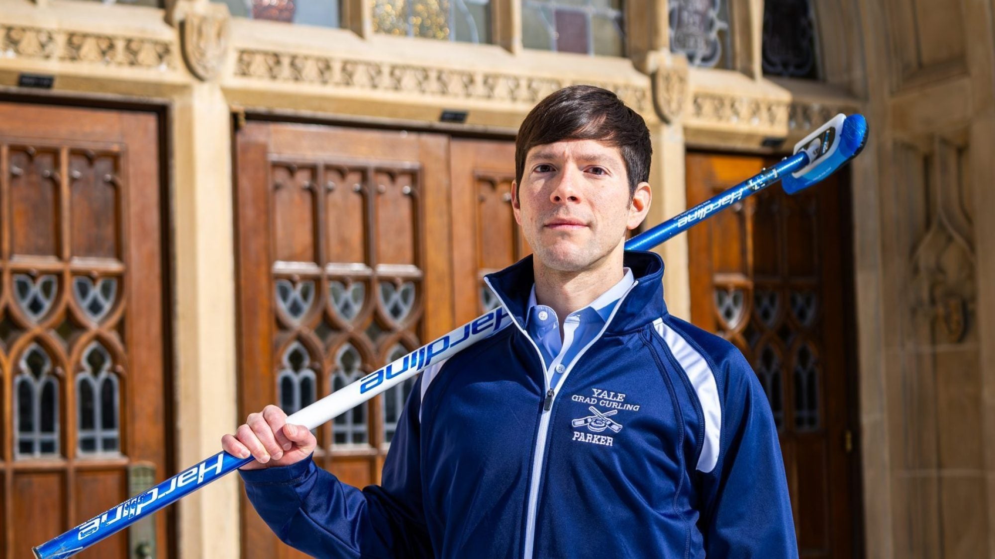 A man with a curling broom stands outside an archway of an academic building