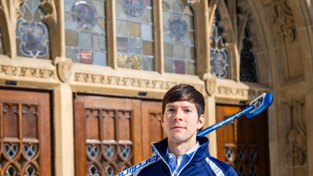 A man with a curling broom stands outside an archway of an academic building