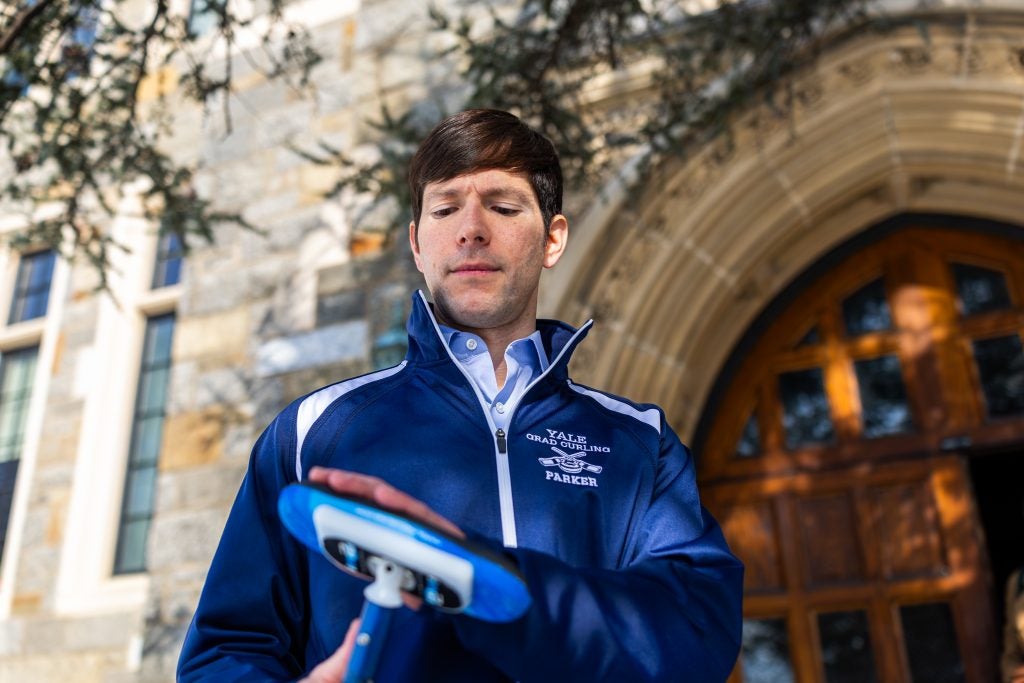 A man in a blue zip-up brushes the top of his curling broom in front of an archway to a building