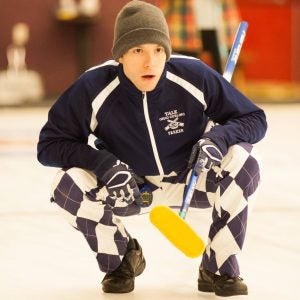 A man in a curling uniform holds a curling broom on ice