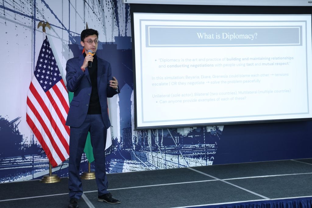 A man in a suit stands in front of an American flag and a screen on a stage