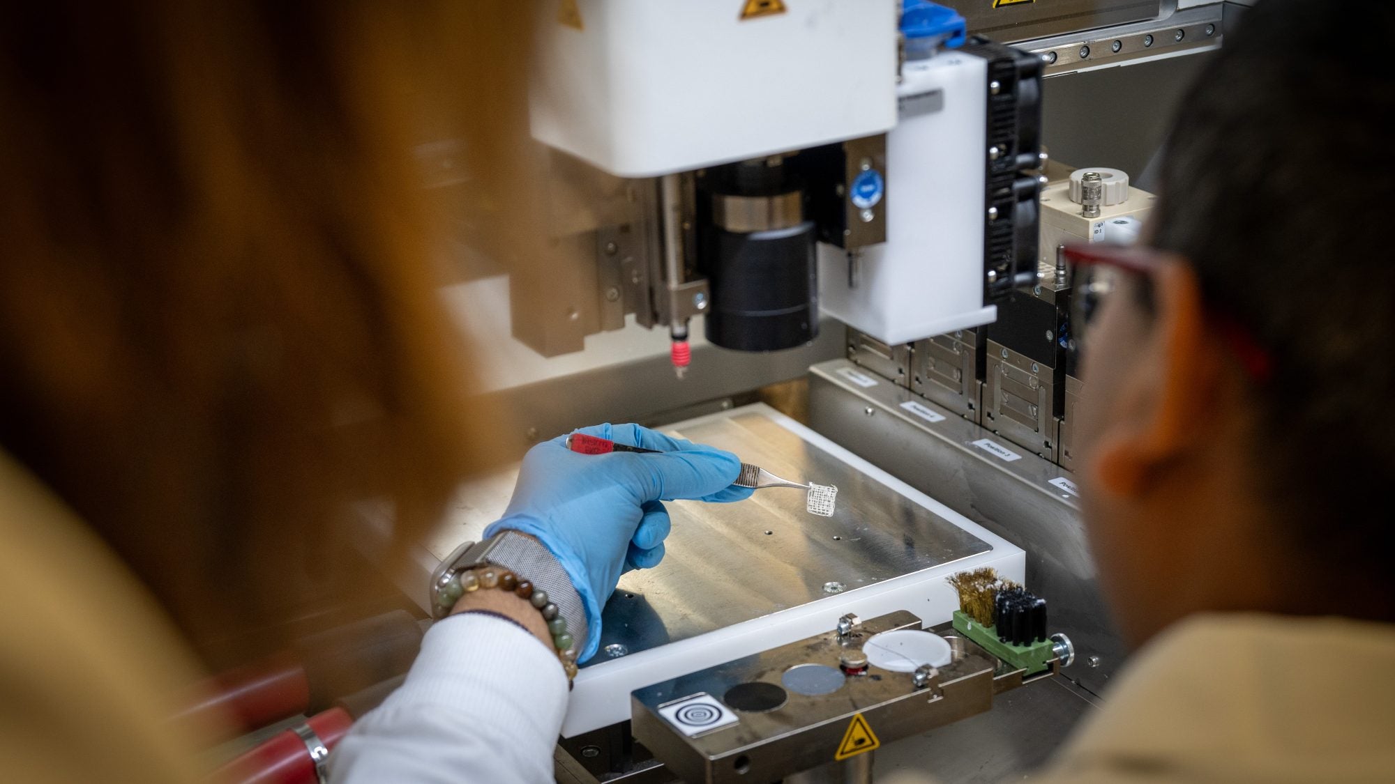 A scientists holds a white square substance with a tweezer by a 3d printer