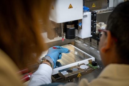 A scientists holds a white square substance with a tweezer by a 3d printer