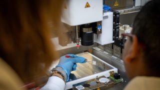 A scientists holds a white square substance with a tweezer by a 3d printer