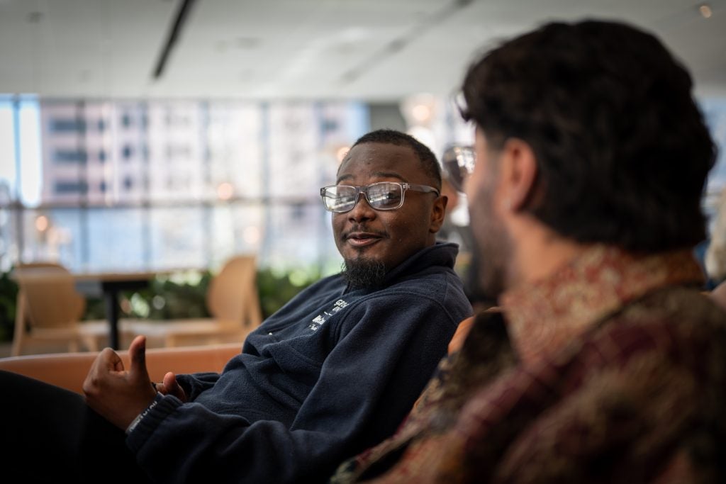 A male student wearing glasses talks to another male student while sitting on a couch