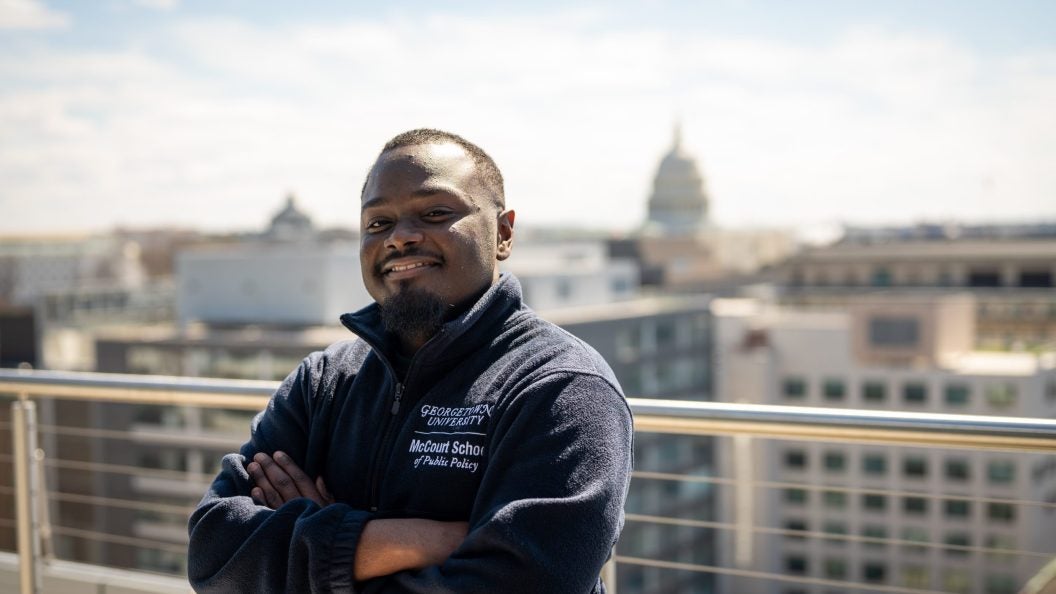 A man smiles with his arms crossed on a balcony. The U.S. Capitol is behind him