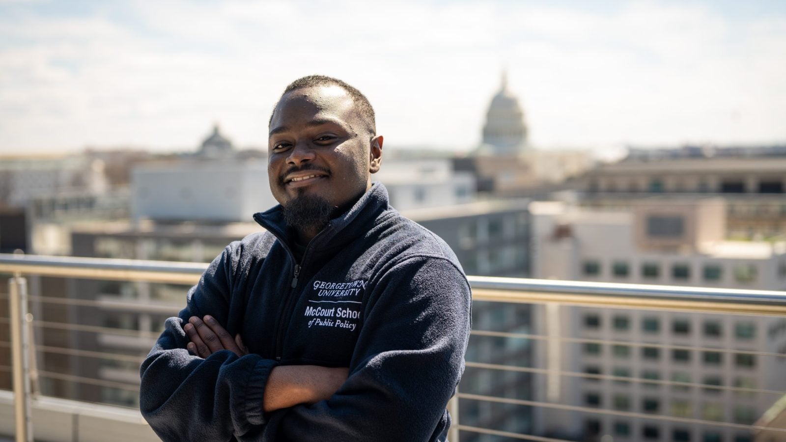 A man smiles with his arms crossed on a balcony. The U.S. Capitol is behind him