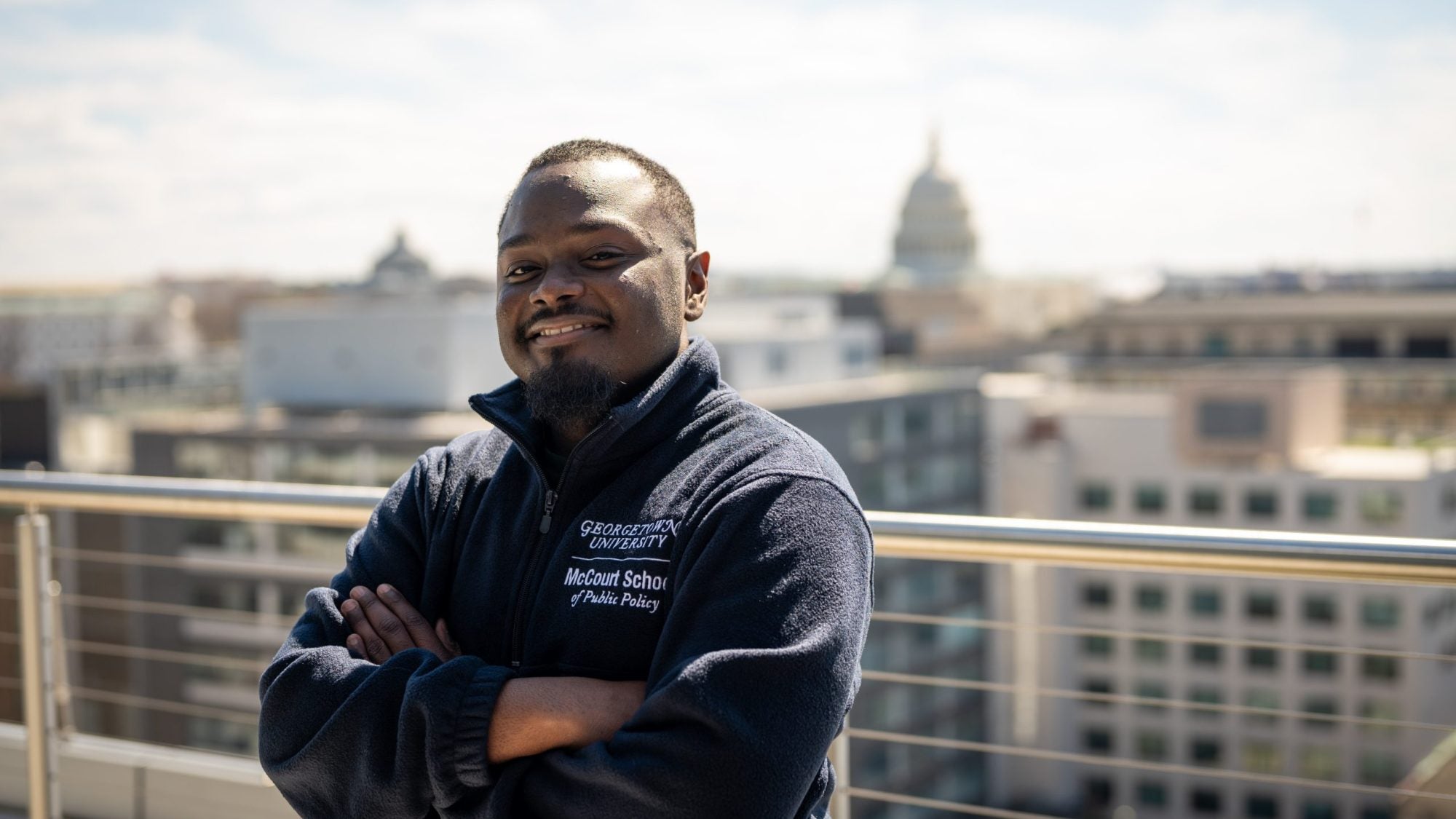 A man smiles with his arms crossed on a balcony. The U.S. Capitol is behind him
