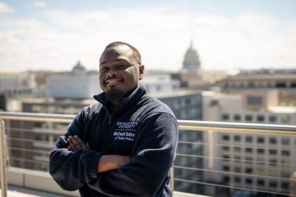 A man smiles with his arms crossed on a balcony. The U.S. Capitol is behind him