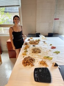 A student stands beside a table of piles of uneaten food and a machine to weigh the amounts