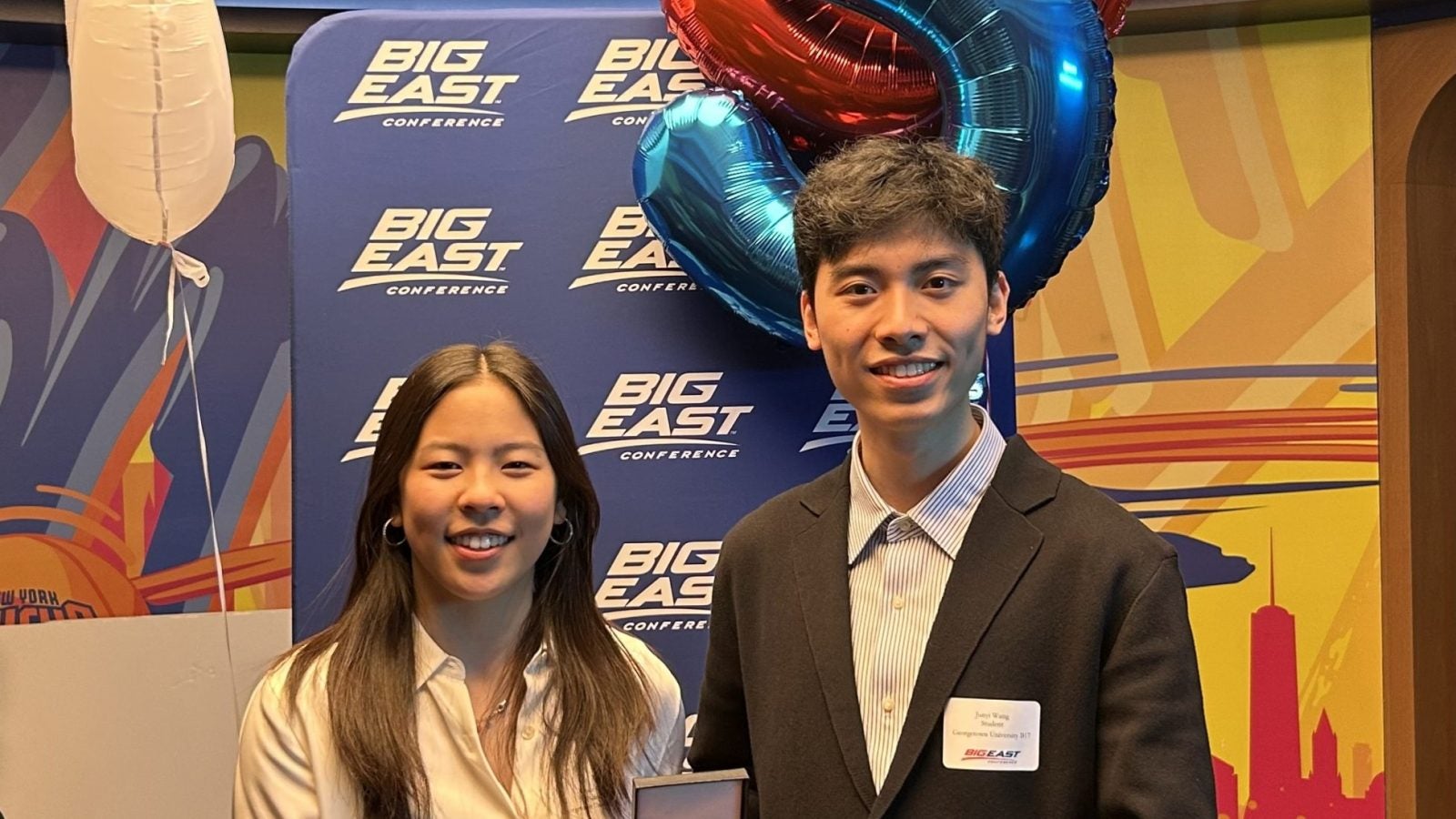 Two students hold a medal in front of balloons of the number "5" and a Big East poster