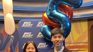 Two students hold a medal in front of balloons of the number "5" and a Big East poster