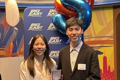 Two students hold a medal in front of balloons of the number "5" and a Big East poster