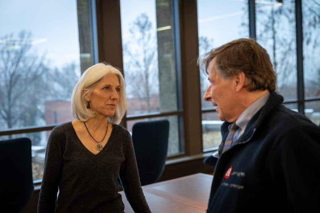 Two people talk to each other in front of large windows