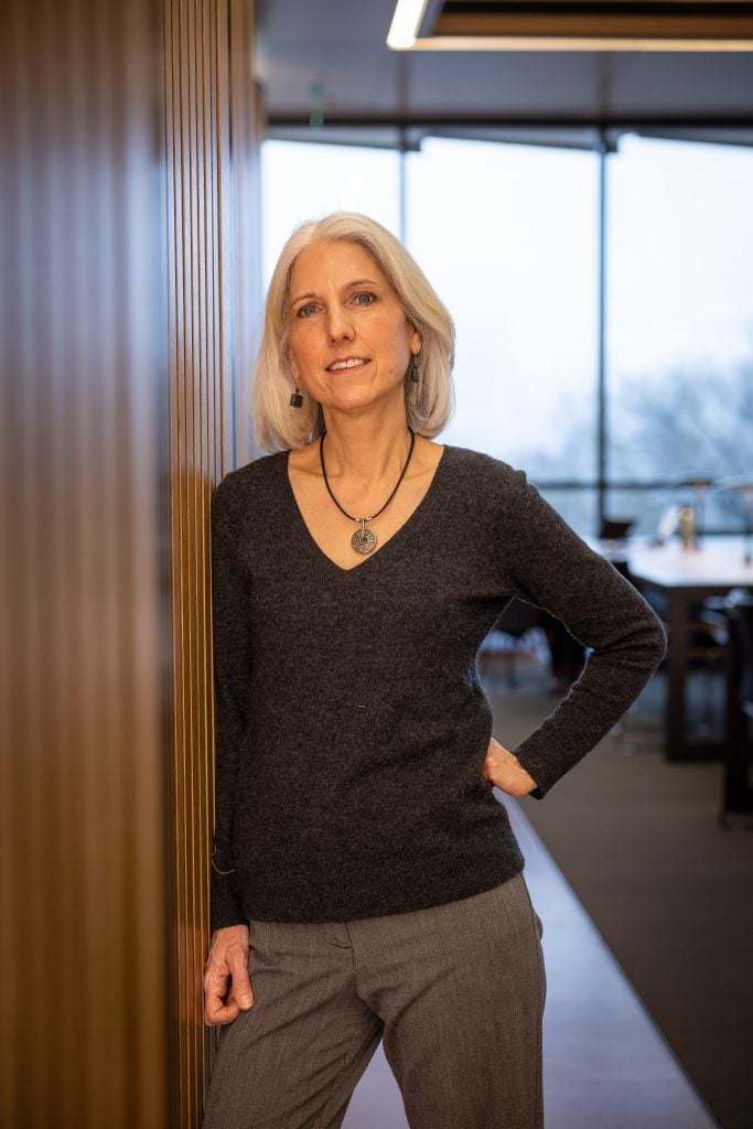 A woman leans against a wall in front of floor to ceiling windows