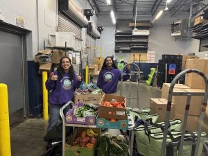 Two women in purple shirts smile at the camera in a food delivery warehouse