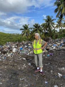 A woman in a neon vest stands in the middle of strewn trash in a landfill. Palm trees are in the background