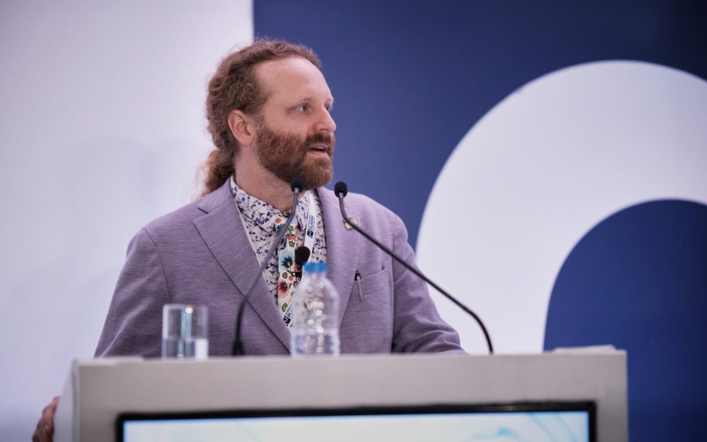A man with red hair and a purple blazer speaks from behind a podium