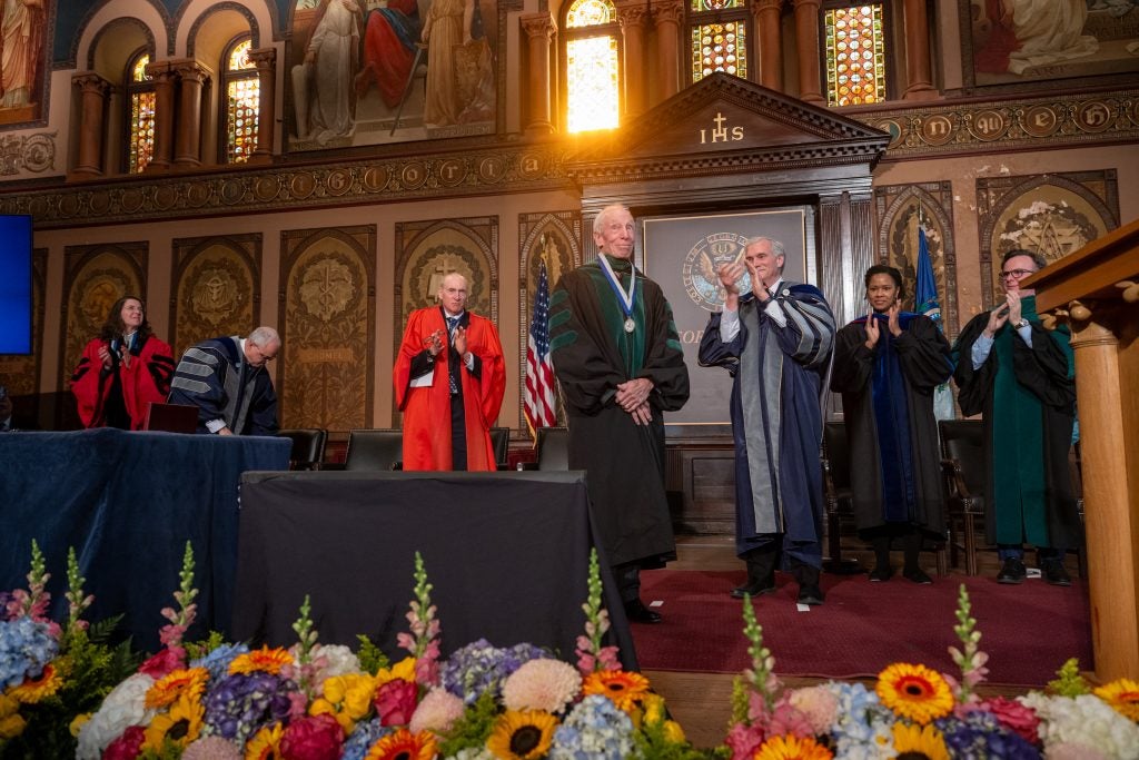 A group of faculty members in academic robes clap for another faculty member dressed in green and black academic robes. Behind him, the sun shines through a stained glass window