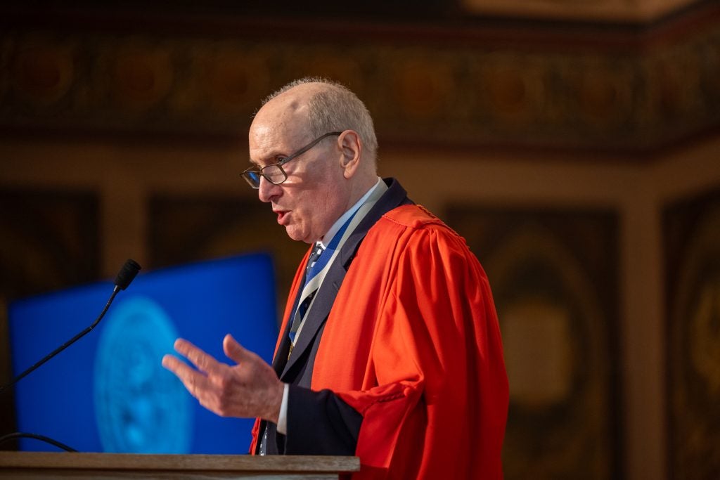 A professor in a red robe gestures while speaking behind a podium