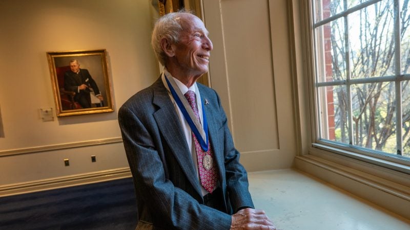 An older man wearing a suit jacket and a medal looks out a window and smiles