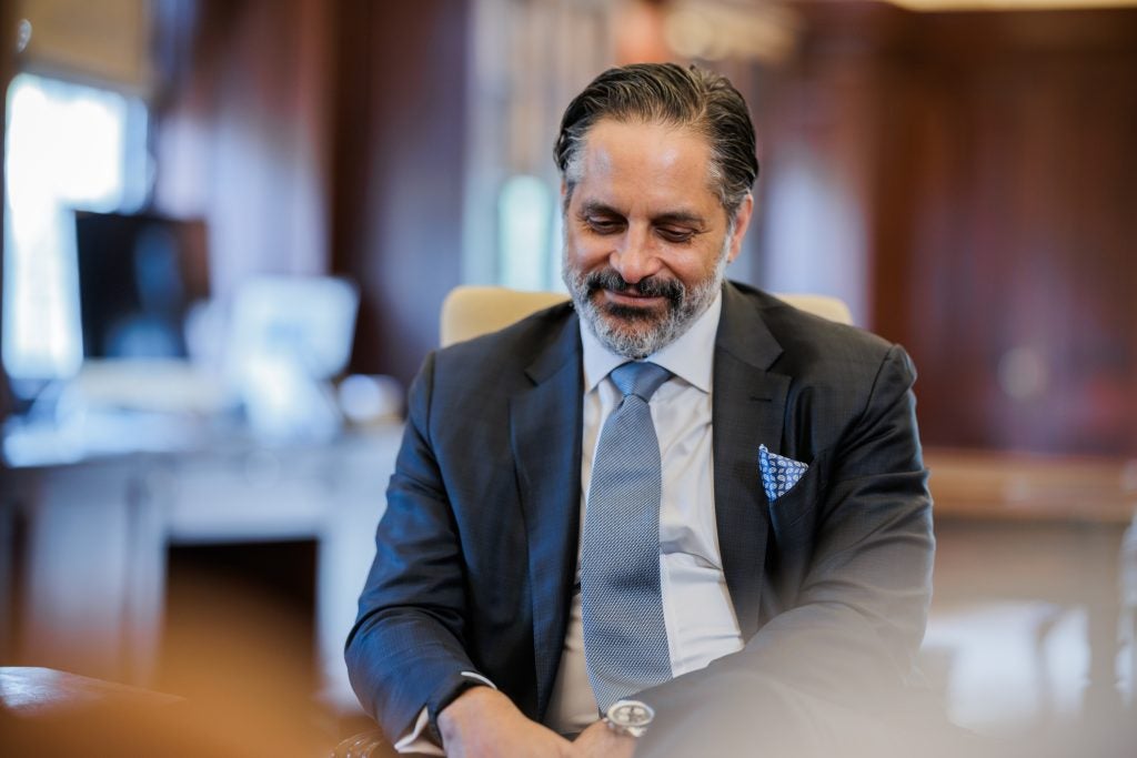 President-elect Eduardo Peñalver smiles and looks down in his office. He wears a dark gray-blue suit and a light blue tie.