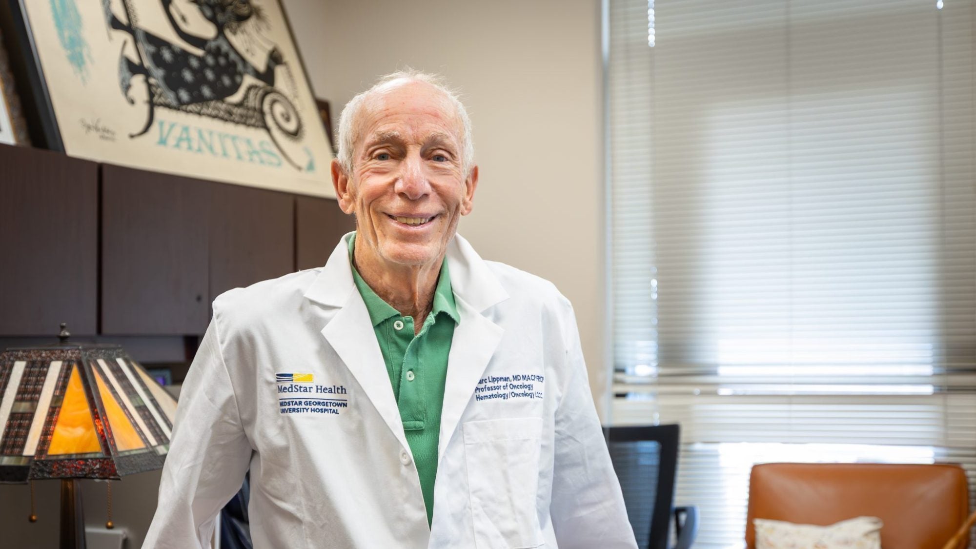 A doctor in a white coat perches on a desk in his office