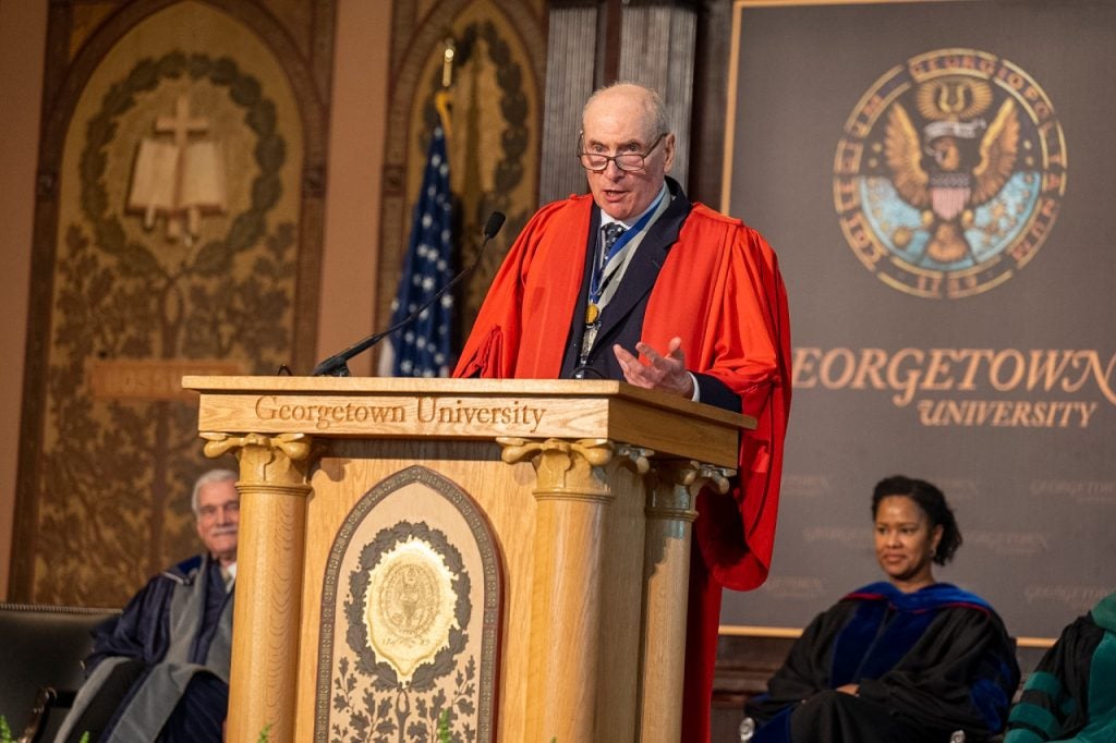 A professor in a red academic robe gestures from behind a podium while speaking on a stage