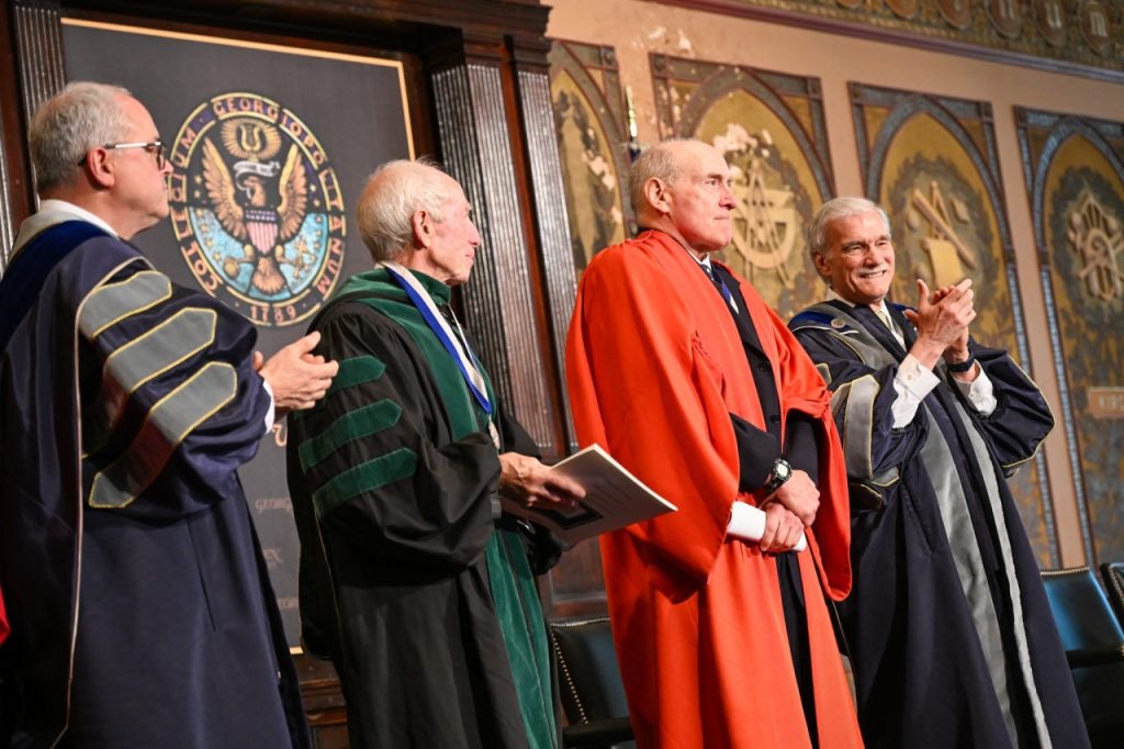 Three males in academic dress robes clap for a professor who's wearing a red academic robe. They all stand onstage with the Georgetown seal behind them.