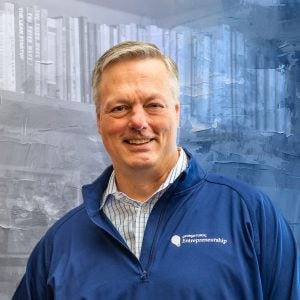 A man in a blue zip-up smiles in front of a textured blue background of books
