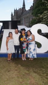A female graduate in a cap and gown poses with her family in front of a sign that says "Hoya Saxa"