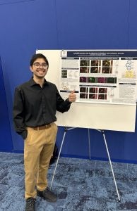 A student smiles and points to a poster with his research on it