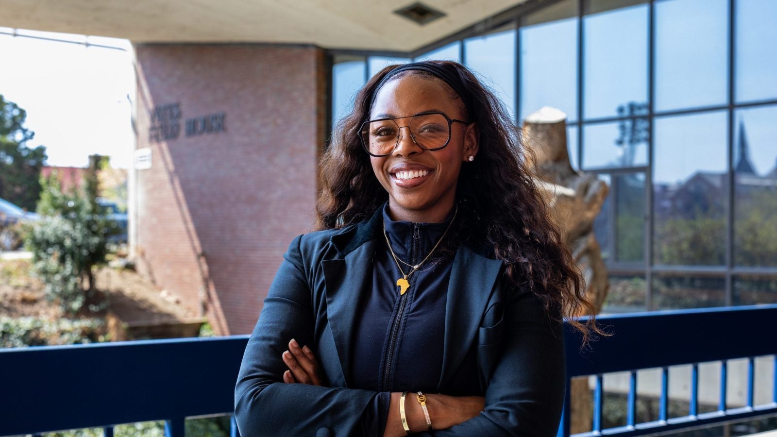 A woman in a blue blazer and clear sunglasses smiles in front of a athletic facility