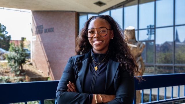 A woman in a blue blazer and clear sunglasses smiles in front of a athletic facility