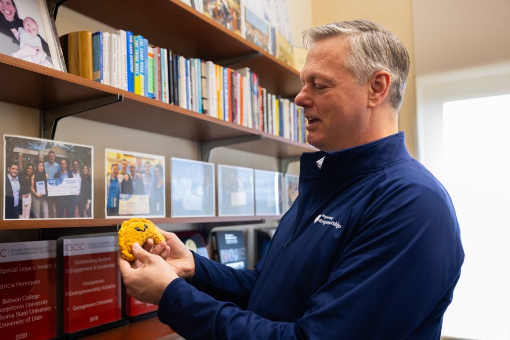 A man in a blue zip-up holds up a knitted croissant toy