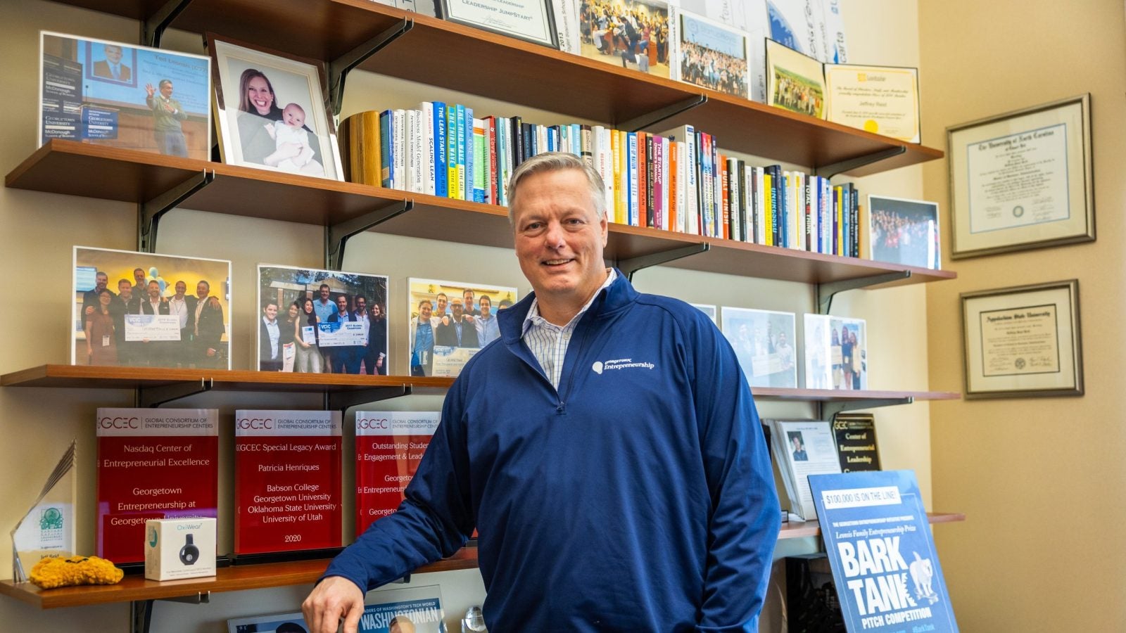 A man in a blue zip-up leans against a bookshelf in his office