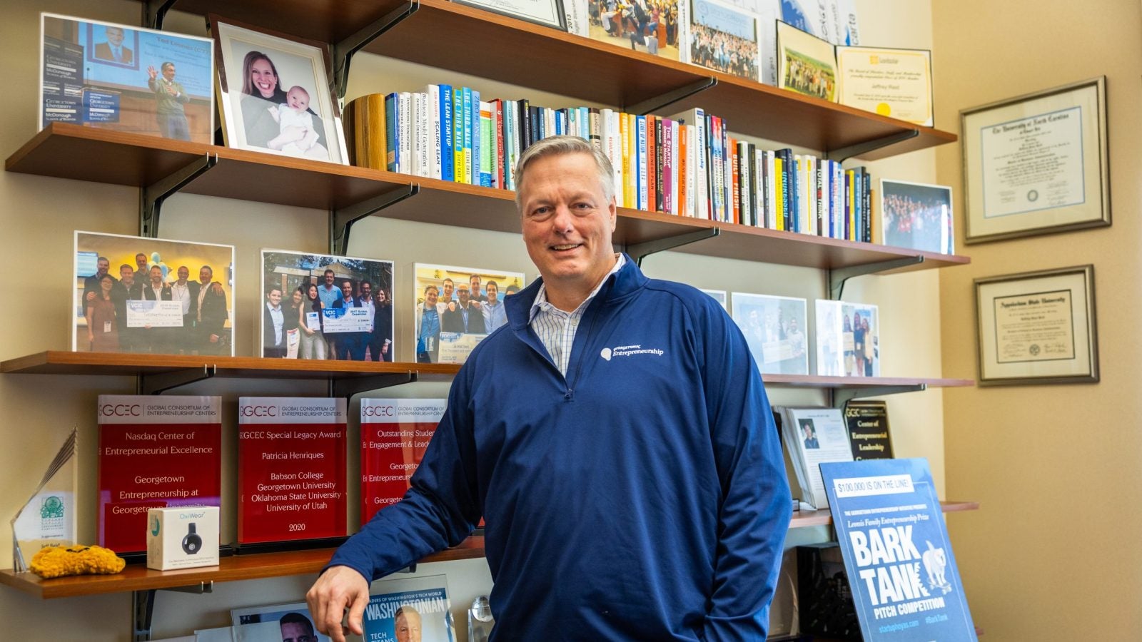 A man in a blue zip-up leans against a bookshelf in his office