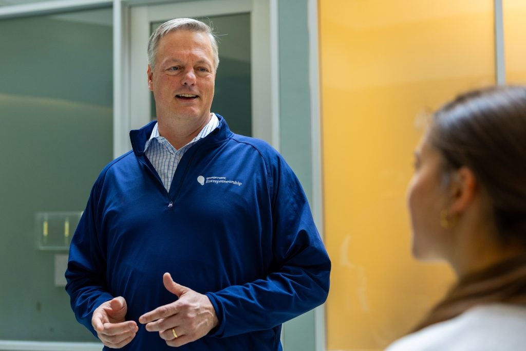 A man in a blue zip-up smiles while talking to a student