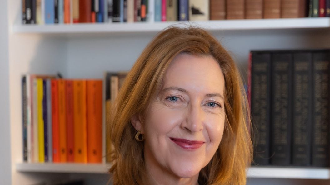 A woman with red hair and a maroon top smiles in front of a bookshelf