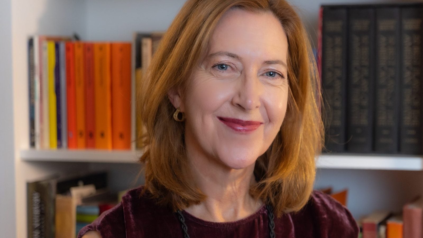 A woman with red hair and a maroon top smiles in front of a bookshelf