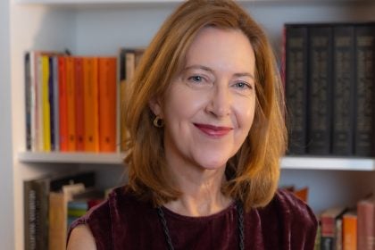 A woman with red hair and a maroon top smiles in front of a bookshelf