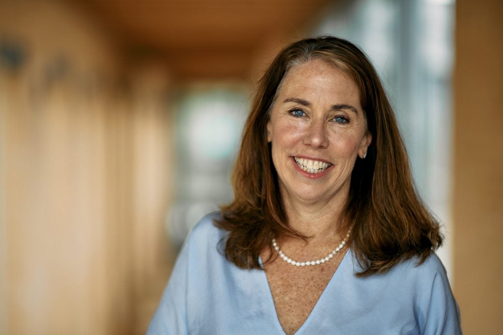 A woman in a blue blouse and pearls smiles in a hallway