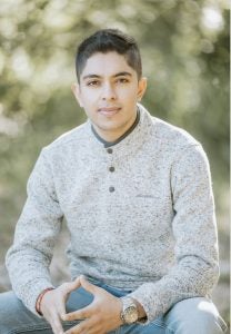 A male student in a gray sweater places his hands together and smiles