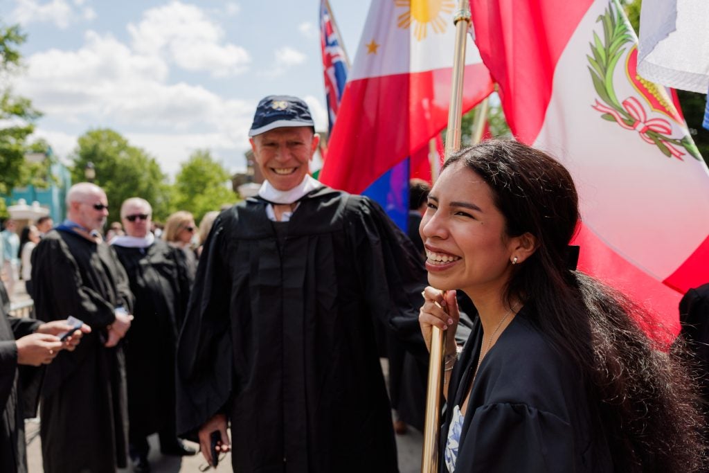 A female graduate holds a flag while smiling and walking in a graduation procession