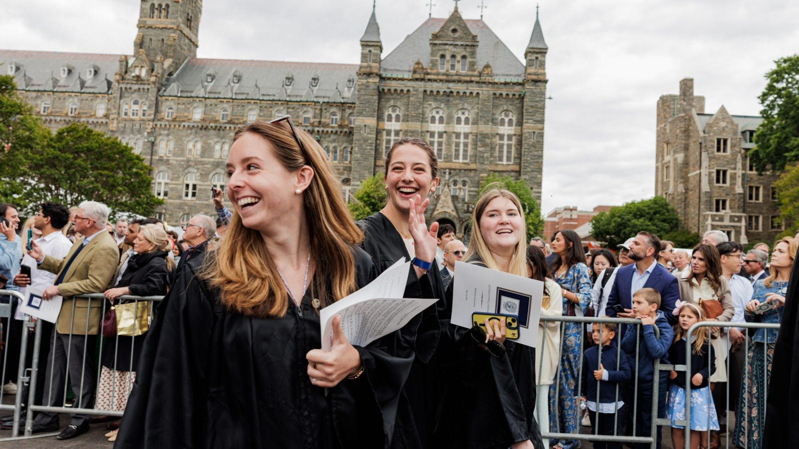 A group of female graduates smile and laugh while walking outside stone buildings on their graduation day