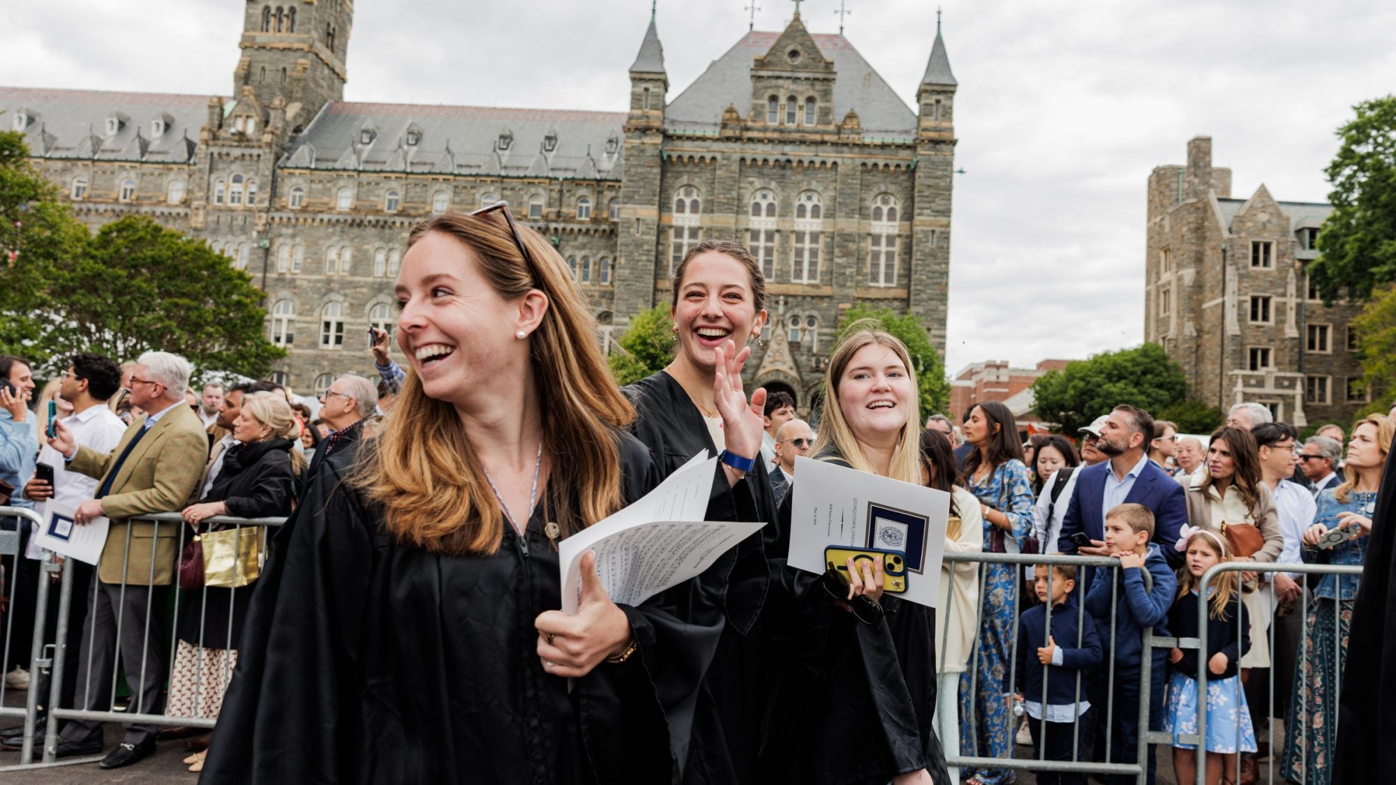 A group of female graduates smile and laugh while walking outside stone buildings on their graduation day