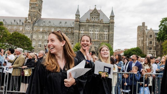 A group of female graduates smile and laugh while walking outside stone buildings on their graduation day