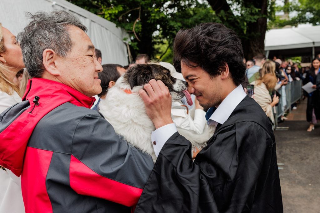 A male student in a black graduation gown cuddles a dog that his relative holds.